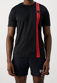 Camiseta deportiva negra con cuello redondo, que presenta dos franjas rojas verticales y un pequeño logo en el pecho. Tejido suave y liviano.