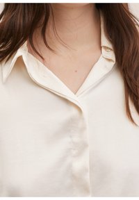 Blouse couleur crème avec une texture douce, dotée d'un col cranté et de boutons sur le devant, soulignant un design classique.