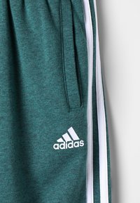 Pantaloni da jogging Adidas color verde acqua con strisce bianche ai lati, tasca con zip e logo Adidas sulla coscia.