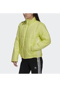 Gul pufferjacka med hög krage, dragkedja fram och vita tre-rands accentueringar på ärmarna. Mjuk textur och gummilogo-detail.