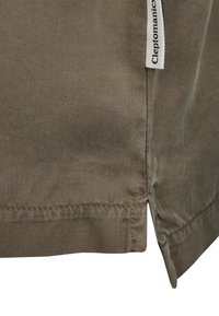 Braunes Baumwollhemd mit einer strukturierten Webart, ausgestattet mit einem seitlichen Schlitz am Saum und einem kleinen weißen Stoffetikett mit dem Markennamen.