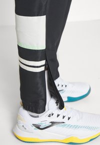 Juodos sportinės kelnių su baltomis ir mėtinėmis detalėmis, su užtrauktuko apykakle, derinamos su baltomis ir mėlynomis sportinėmis batelėmis su geltonu padu.