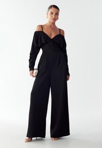 Moteris juodu off-shoulder šokiu su plačiomis kelnėmis, laikanti mažą metalinę rankinę ir dėvinti auksines auskarus-žiedus bei sandalinius batus su juostelėmis.