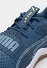 Blå atletisk sko med texturerade mesh-paneler, gummisula, tjocka skosnören och en framträdande logotyp. Accentdetaljer i ljusblått.