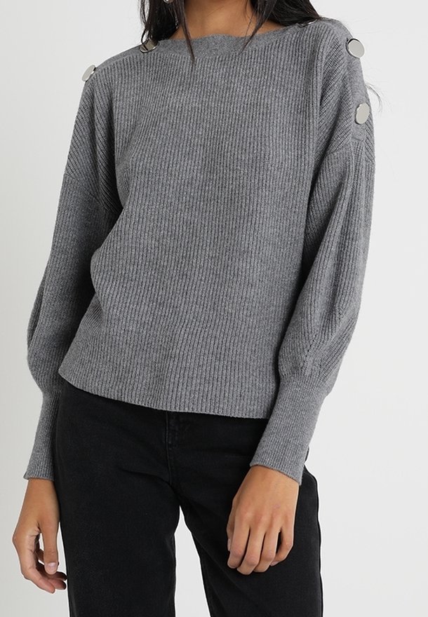 Grijze geribbelde trui met een brede halslijn en ballonmouwen, voorzien van metalen knoopaccenten op de schouders. Gecombineerd met zwarte broek.