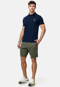 Polo marine avec col, manches courtes et détail de logo ; associé à un short vert olive et des chaussures de sport blanches avec des accents.