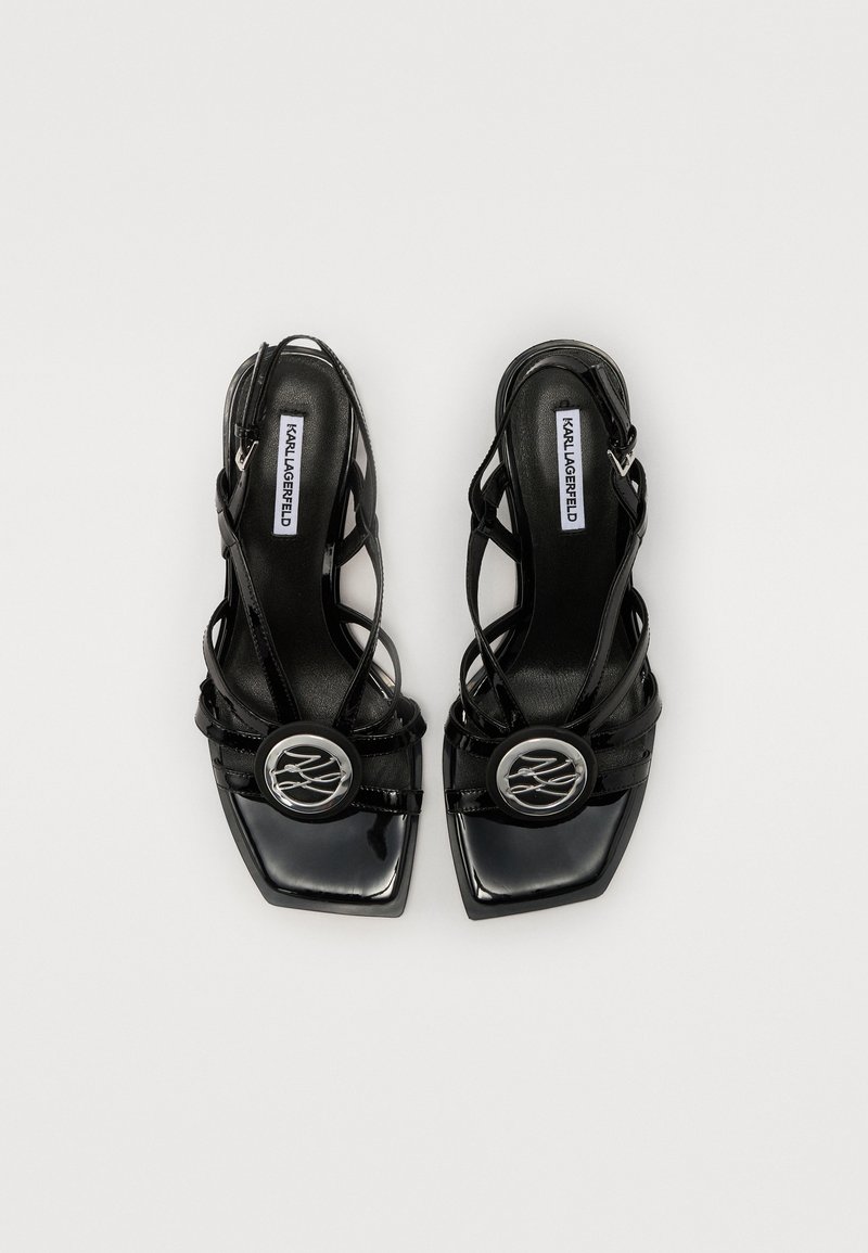 Sandales à talons en cuir verni noir avec un embout carré, des lanières croisées et un détail en logo argenté circulaire. Bride de cheville réglable incluse.