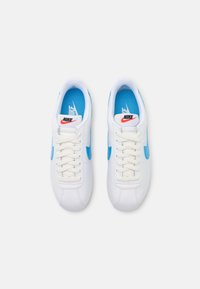 Valged Nike tossud siniste aktsentidega, millel on tekstureeritud ülemine osa, ümar varvas, lamedad paelad ning logo keele ja külje peal.