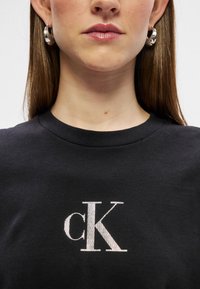 Crna pamučna majica s bijelim vezenim "cK" logotipom. Ovratnik je klasičnog rebrastog kružnog stila, s glatkom teksturom.