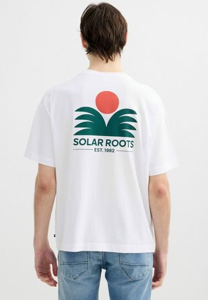 Persona con cabello corto y marrón que lleva una camiseta blanca con un logo de una hoja verde y un sol rojo y el texto "SOLAR ROOTS EST. 1982" en la espalda.