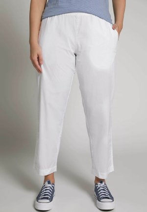 Widok osoby od talii do stóp w białych spodniach z elastycznym pasem, z rękami w kieszeniach i niebieskich płóciennych trampkach na jasnym tle.