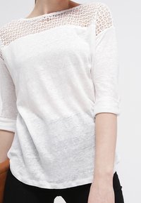 Bílý dlouhý rukáv top s texturovaným krajkovým yoke, měkkou tkaninou, kulatým výstřihem a ohrnutými manžetami, spárovaný s černými kalhotami.