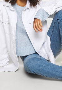 Weißes Hemd mit Knöpfen, das über einem hellblauen Strickpullover mit gerippten Bündchen getragen wird. Hellblaue Jeans mit einer körpernahen Silhouette.