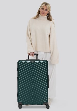 Donna con un maglione beige e pantaloni bianchi tiene il manico di una valigia rigida verde scuro con motivo a zigzag e quattro ruote.