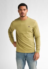 Sudadera verde oliva con escote redondo y mangas largas. Presenta un pequeño logo en el pecho y un dobladillo ajustado. El material parece suave.