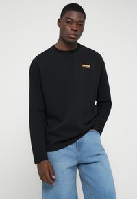 Tricou negru cu mânecă lungă din bumbac, având un logo portocaliu "hummel" pe piept. Asortat cu blugi din denim de culoare albastră deschisă.