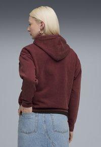 En blond person iført sølvfarvede hoops øreringe, en maroon hættetrøje og en lyseblå denim nederdel med den ene hånd i baglommen, vendt mod venstre.