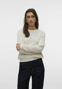 Sweter w kolorze kremowym, z dzianiny, z teksturowanym wzorem, z prążkowanymi mankietami i dołem. Zestawiony z ciemnoniebieskimi dżinsami. Prosty, casualowy fason.