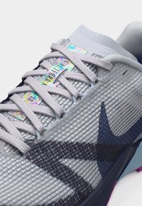 Αθλητικό παπούτσι ανοιχτού γκρι με υφή στο πάνω μέρος, μπλε σκούρο λογότυπο Nike, ολογραφική ετικέτα "Nike Terra Kiger 10" και κορδόνια στο ίδιο γκρι χρώμα.