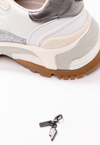Gros plan sur un talon de sneaker blanc et argenté avec une semelle texturée et un petit pendentif en métal noir en forme d’éclair sur une surface blanche.