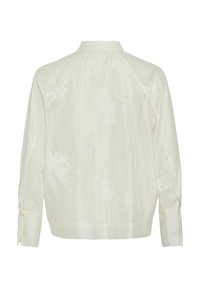 Blouse blanche brodée à manches longues et col. Présente des motifs floraux et un tissu texturé avec une légère brillance.