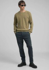 Olijfgroene gebreide trui met geribbelde manchetten en zoom, gecombineerd met donkergrijze broek en zwarte enkellaarzen, model draagt zwarte zonnebril.