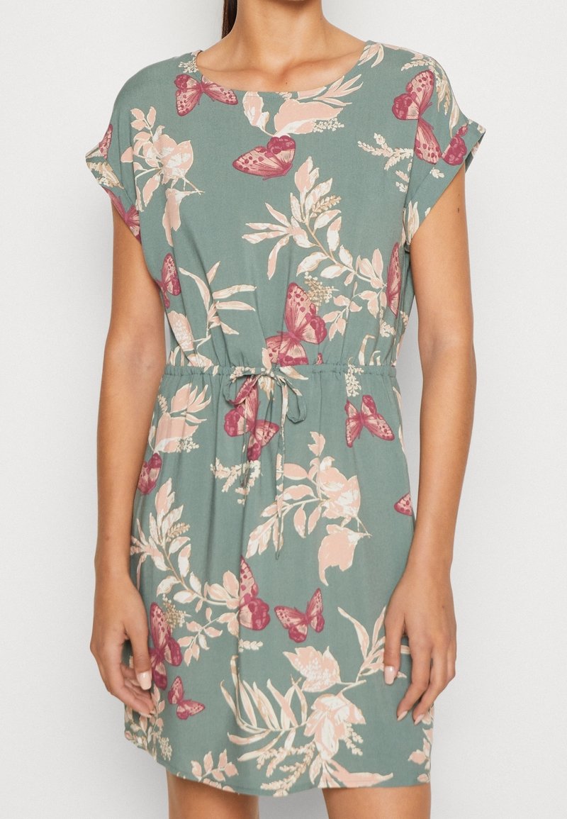 Robe courte à manches courtes verte avec motif papillon rose et motifs floraux beige, taille élastique avec nœud à l'avant, portée par une femme, vue du cou jusqu'à la mi-cuisse.