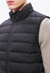 Μαύρο αμάνικο μπουφάν με ψηλό γιακά, ραμμένο σχέδιο και κλείσιμο με φερμουάρ. Διαθέτει διακριτικό λογότυπο στο στήθος και υφασμάτινη υφή.