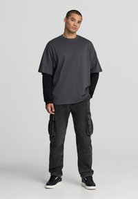 T-shirt grigia oversize sovrapposta a una maglietta a maniche lunghe nera, abbinata a pantaloni cargo neri con molte tasche e sneakers nere.