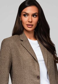 Hnědý herringbone blazer s jedním knoflíkem na zapínání, sklopenými klopami a klopovými kapsami, nošený přes bílé tričko. Hladká textura.
