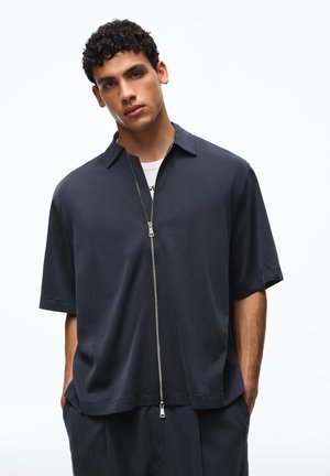 Young man with curly hair wearing a dark navy short-sleeve zip-up shirt and matching pants, standing with hands in pockets.