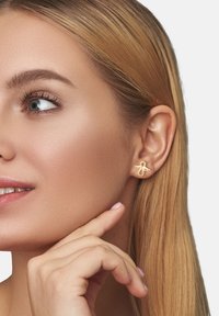 Goldene Metallohrringe, die wie ein kleiner Schmetterling geformt sind, mit glatter Oberfläche und subtilen Details, getragen von einem Modell mit langen Haaren.