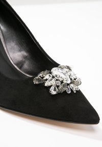 Escarpin en daim noir à bout pointu orné d'un groupe de strass clairs de formes diverses près de la zone des orteils.