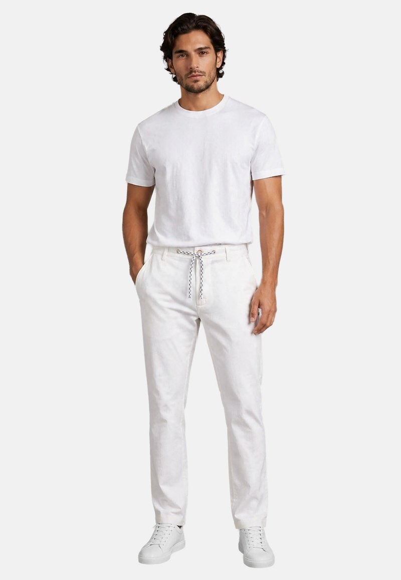Mann mit dunklen Haaren trägt ein komplett weißes Outfit: kurzärmeliges T-Shirt, Hose mit Kordelzug und weiße Sneakers, steht vor einem einfarbigen Hintergrund.