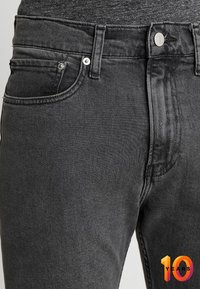 Los jeans de mezclilla negros tienen un ajuste slim, un diseño clásico de cinco bolsillos y detalles en metal en los botones y remaches. La tela presenta una ligera textura.