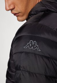 Giacca a vento nera con design trapuntato e cuciture orizzontali. Presenta un logo Kappa ricamato sulla spalla in grigio chiaro.