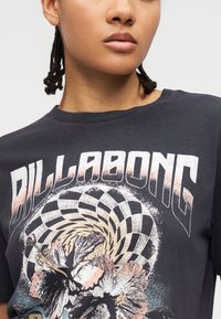 Άτομο που φοράει σκούρο μπλουζάκι Billabong με σπειροειδές καρό μοτίβο και μεγάλο λουλουδάτο σχέδιο σε παστέλ αποχρώσεις.