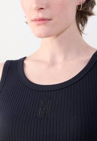 Canottiera nera a costine con scollatura rotonda e un logo ricamato discreto sul davanti. Il tessuto ha una texture liscia.