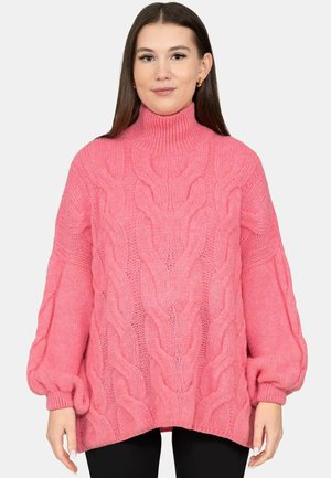 Frau trägt einen locker sitzenden pinkfarbenen, grob gestrickten Rollkragenpullover mit Ballonärmeln, stehend vor schlichtem hellem Hintergrund.