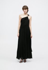 Vestido longo preto plissado com design de um ombro só e detalhe de fita. Tecido leve que cai elegantemente até ao chão. Saltos pretos com tiras.