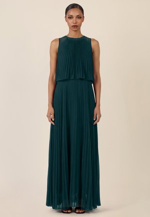 Vrouw draagt een mouwloze, donkergroene, geplooide jurk tot op de vloer en staat tegen een effen beige achtergrond.
