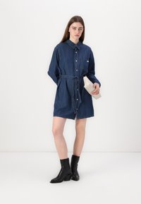 Abito camicia in denim di colore blu scuro, con colletto, chiusura con bottoni davanti e cintura in vita. Abbinato a stivaletti neri e una pochette color crema.