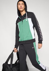 Μαύρο, πράσινο και λευκό φόρμα με φούτερ με φερμουάρ. Διαθέτει λογότυπα, ριγέ πλευρές και άνετη εφαρμογή. Περιλαμβάνει μαύρη τσάντα γυμναστηρίου.