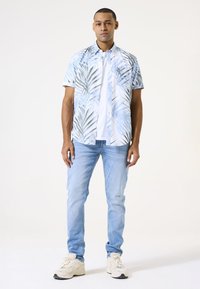Camisa de manga corta abotonada con un patrón de hojas de palma azul y blanco; combinada con jeans de color azul claro y zapatillas beige.