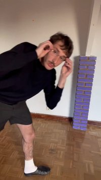 Homme ajustant ses lunettes en se penchant près d'une haute pile de briques violettes à l'intérieur, sur un sol en parquet.
