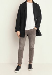 Svart långärmad kappa över en vit t-shirt, kombinerad med ljusgrå jeans och svarta sneakers. Kappan har en strukturerad design och knappdetaljer.