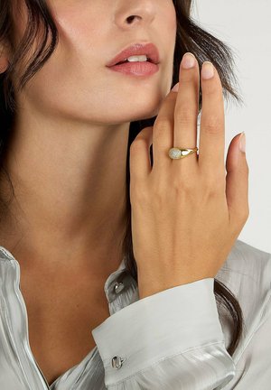 Frau trägt eine blasssilberne Bluse und hebt die Hand mit einem goldenen Ring, der einen runden, mit Diamanten besetzten Cluster auf ihrem Ringfinger zeigt, in der Nähe ihres Gesichts.