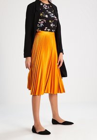 Plisowana pomarańczowa spódnica z błyszczącego materiału, zestawiona z czarną kwiecistą bluzką i czarną kurtką; czarne płaskie buty dopełniają stylizację.
