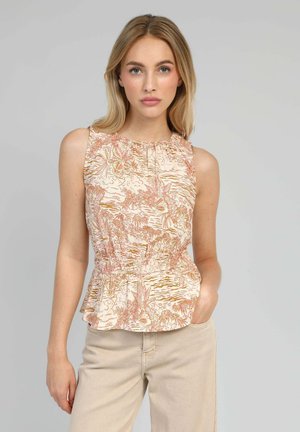 Jeune femme portant un haut sans manches beige à fleurs avec une taille smockée, debout avec une main dans la poche sur un fond gris uni.