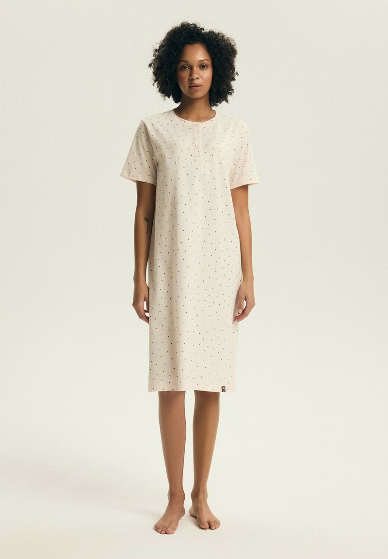 Žena stojaca bosá, oblečená v krémových šatách po kolená s krátkym rukávom a malými čiernymi bodkami na jednoduchom svetlom pozadí.
